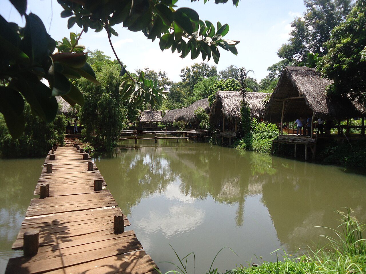 Thatched roofs on stilts above a pond at Ako Dhong, with a wooden rail in front