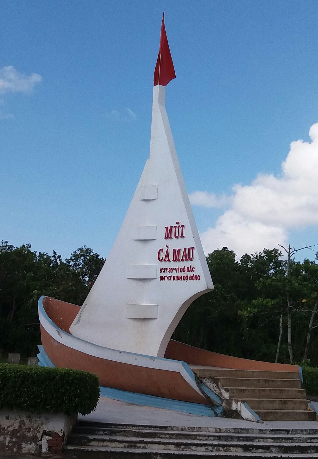 White sail monument reading Mũi Cà Mau, with a red flag, steps, and mangrove trees
