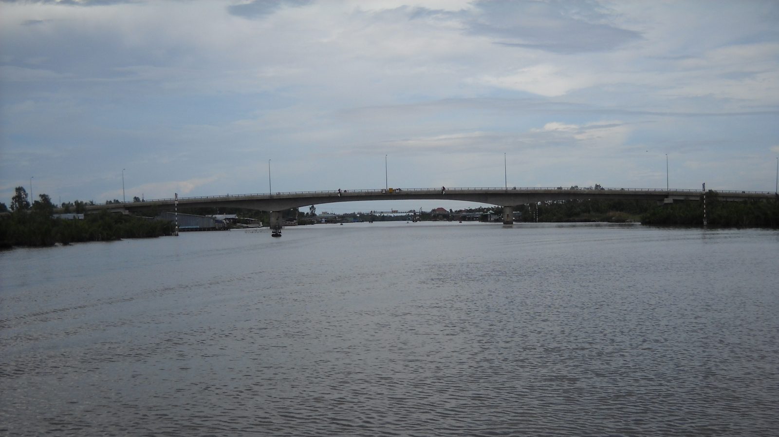 A long concrete bridge across the wide Ông Đốc river, with a small boat underneath