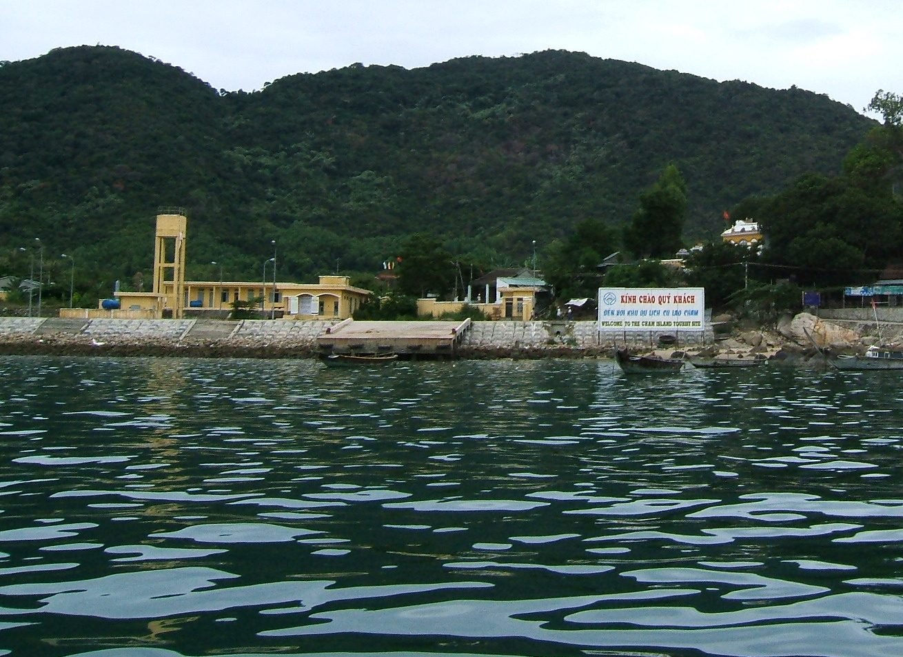 The Bai Lang shore and a small pier, seen from the water, with a yellow tower and forested hill