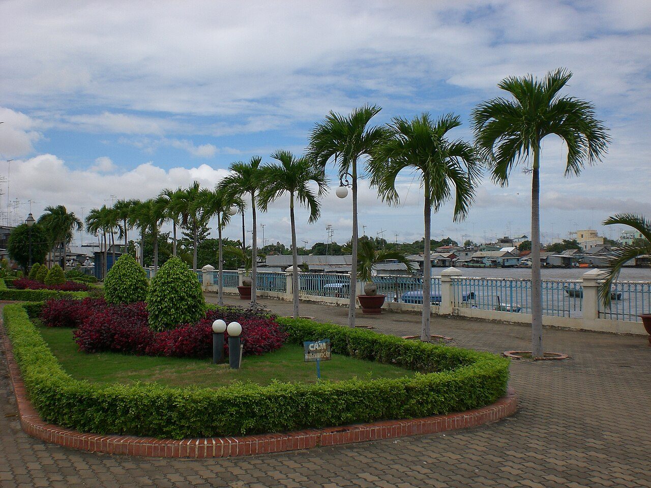 A palm-lined riverside walk in Chau Doc, with the river and houses beyond the railing