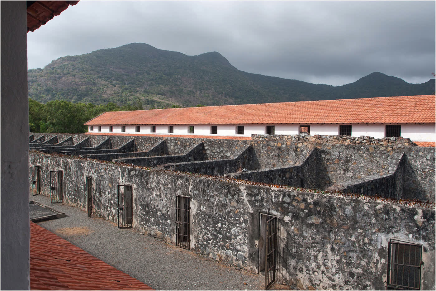 Stone prison walls with barred doors and a long cell block under a tiled roof, with the ridge behind