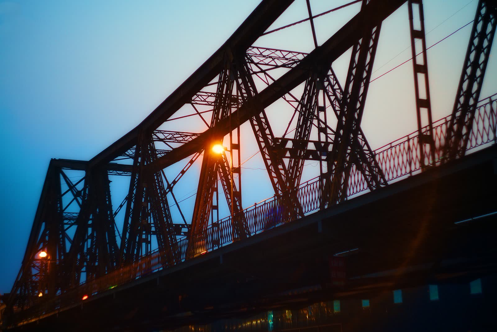 The riveted steel lattice of Long Bien Bridge at dusk, with a lamp lighting the span