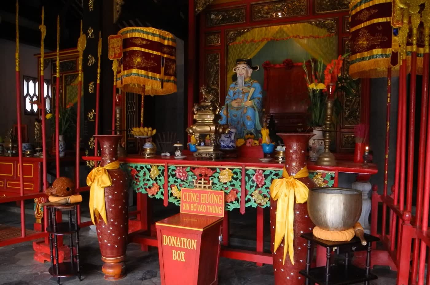 A seated statue in a blue robe inside Quan Cong Temple in Hoi An, with an incense burner and red banners
