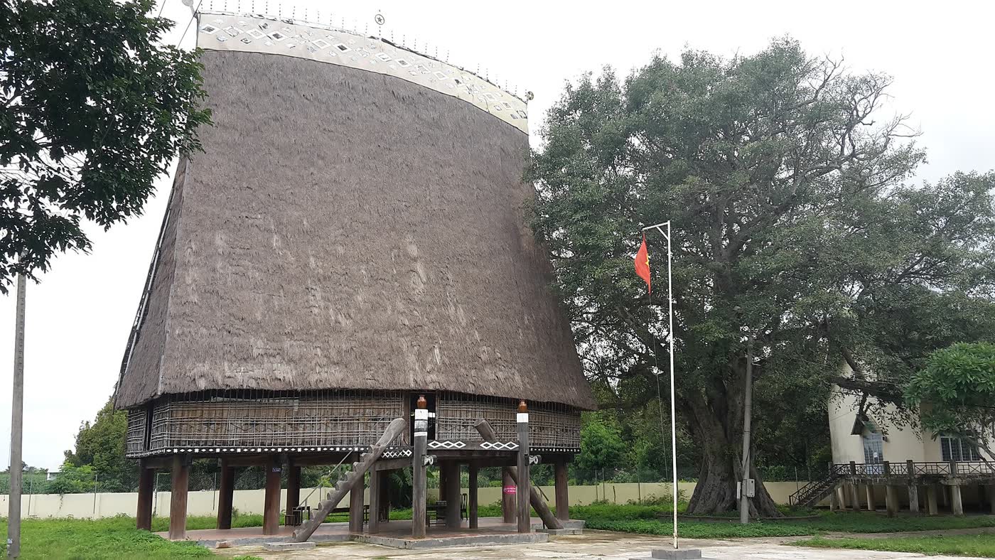 The Kon Klor communal house from the side, thatch roof and stilts, with a large tree in the yard