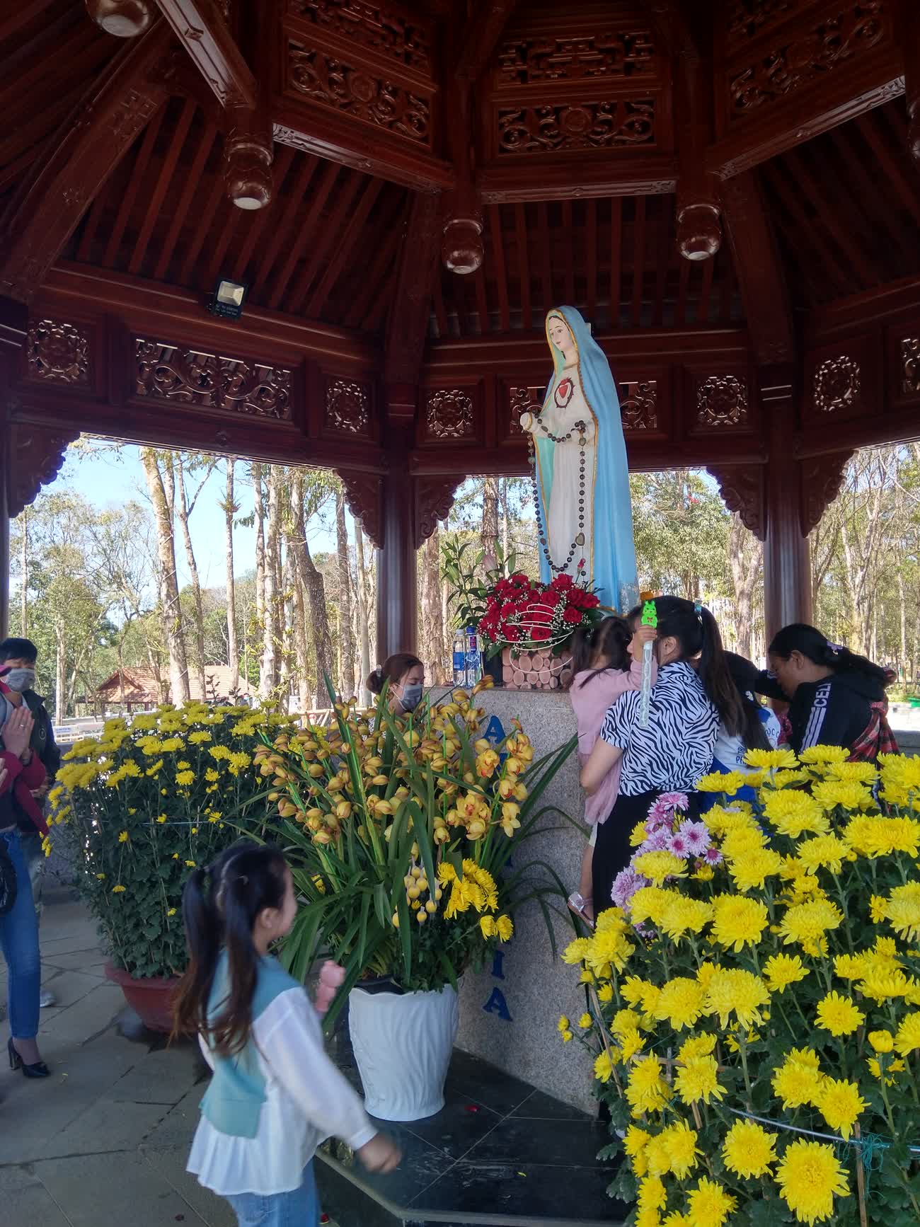 The Virgin Mary statue inside a wooden pavilion, with flowers and visitors at the base