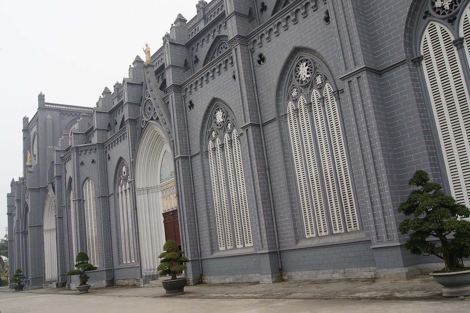 The side of Phu Nhai basilica, gray stone and tall arched windows, with potted trees