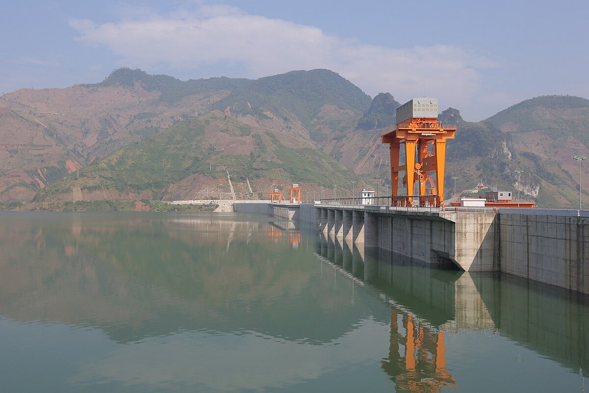 The Son La Dam from the reservoir, with the concrete crest, an orange gate, and forested hills